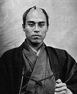 Portrait of Fukuzawa Yukichi sitting. He is a middle aged man with a balding head. He is wearing a philosophers robe and is staring intently at the camera. 