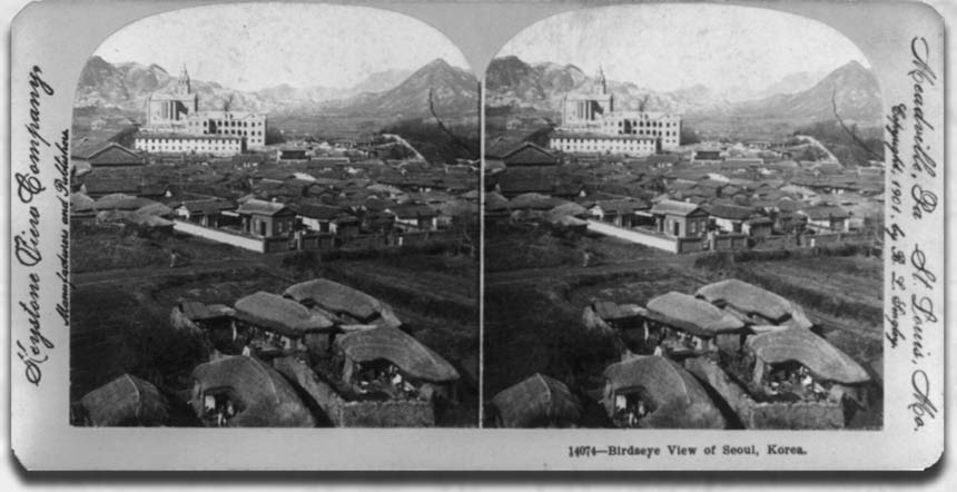 aerial view of a village in front of a large stately building. in the distance are mountains.