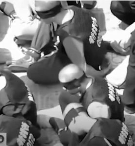 aerial photo of people sitting on the floor with their hands tired behind their backs. they are blindfolded and their feet are chained, and their heads are shaved bald.