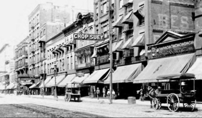 An old picture of the street life in early-20th Century Chicago. The buildings are all three to five floors high, all with a canopy outside. Several coaches are on the street. A banner writing "Chop Suey" is hanging on one building. 
