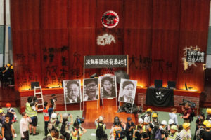 photograph of a hall with several photos and graffiti written on the wall. many people in hard hats and reflective wear stand in front.
