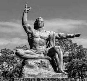 a statue of a man sitting on a boulder, one leg crossed under him, the other leg sitting forward. his left hand points upwards, while he uses his right hand, palm facing down, to point out and away from his body.