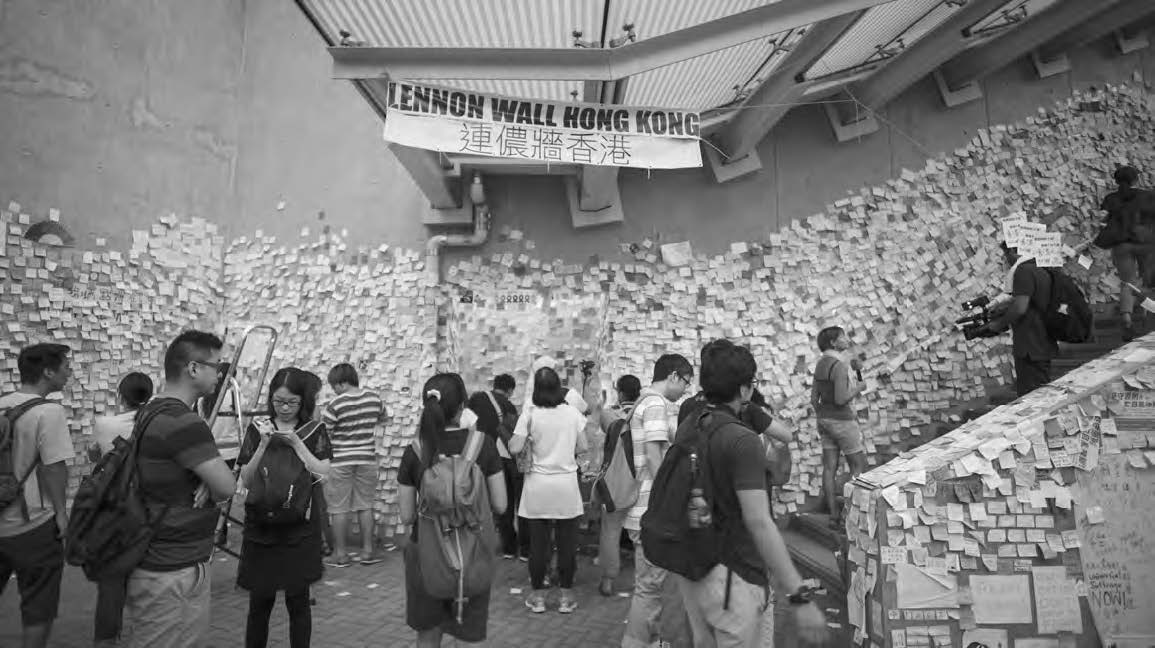 many people stand in front of the "lennon wall hong kong" which is covered in notes