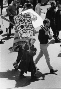black and white photo of protestors. a young person holds a sign that says "bring the troops home now"