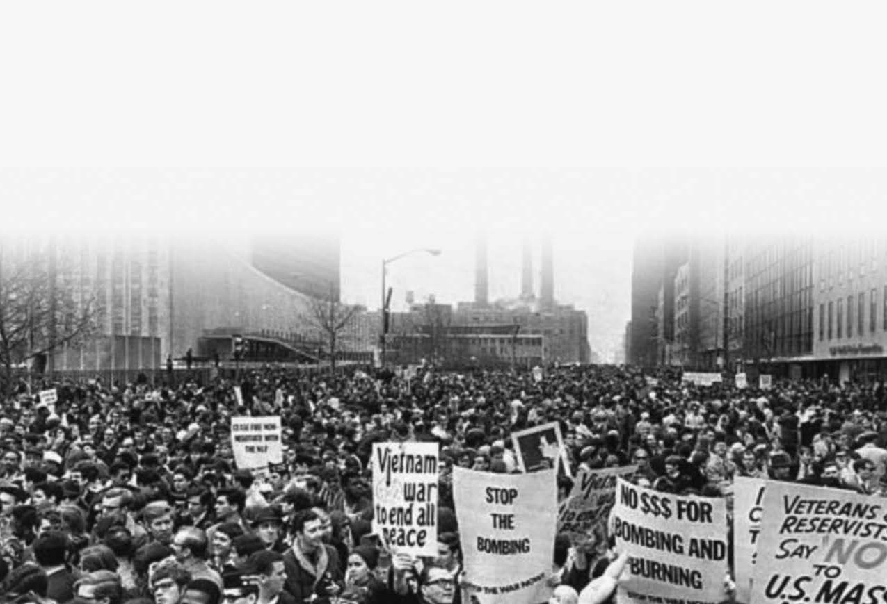 photo of a large group of people protesting white holding protest signs saying "no $$$ for bombing and burning" and "vietnam war to end all peace"