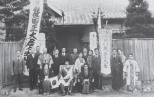 black and white photo of a group of people in suits, kimono, and military uniforms with banners written in japanese on either side of them