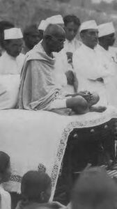 a man sits crossed legged on a podium as others dressed in white stand behind him