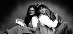 photo of a man holding an instrument sitting back to back with a woman in glasses and holding a book