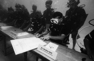 the image in underwater, with several people in scuba suits standing at a long table to sign government documents